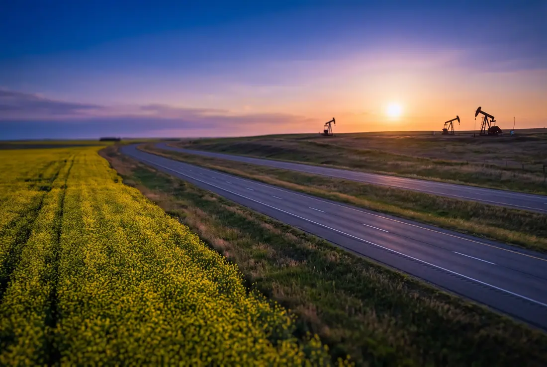 Divided prairie highway at sunset beside a yellow canola field, pump jacks on the horizon